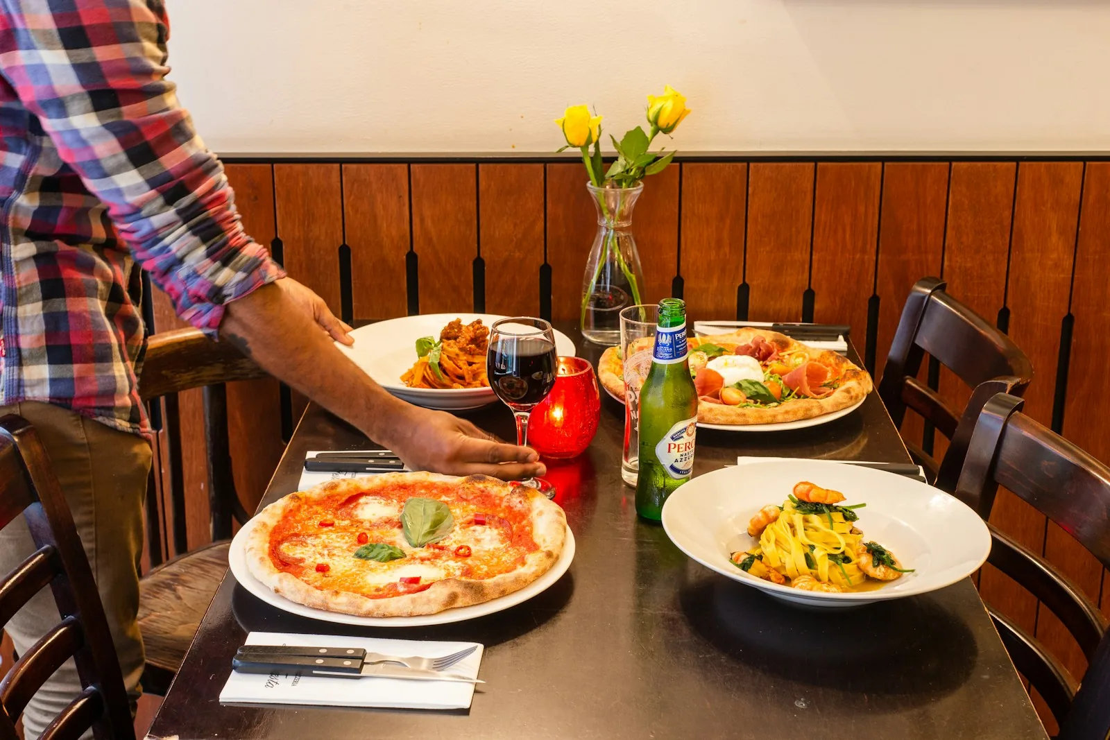 A server placing plates of pizza and pasta on a wooden restaurant table set with candles and wine glasses — the operational reality the manager is responsible for getting right, every cover, every shift